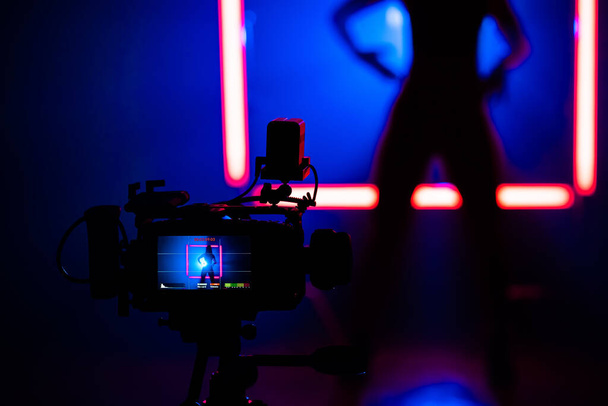 Backstage how operator working with professional camera. Videographer shoots clip with sexy woman dancing in dark studio with neon color light lamps. - Φωτογραφία, εικόνα