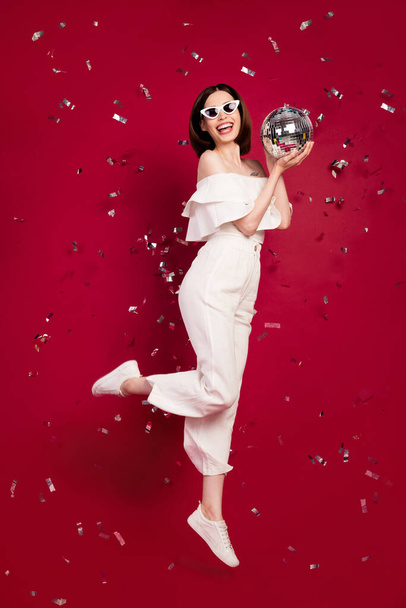 Vertical full length body size photo pretty brunette keeping discoball fumping flying confetti isolated red color background - Foto, afbeelding