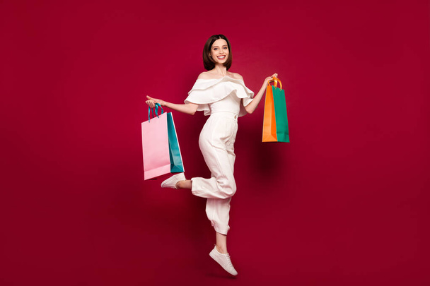 Full length body size photo pretty brunette jumping up smiling keeping bags after shopping isolated red color background - Fotoğraf, Görsel