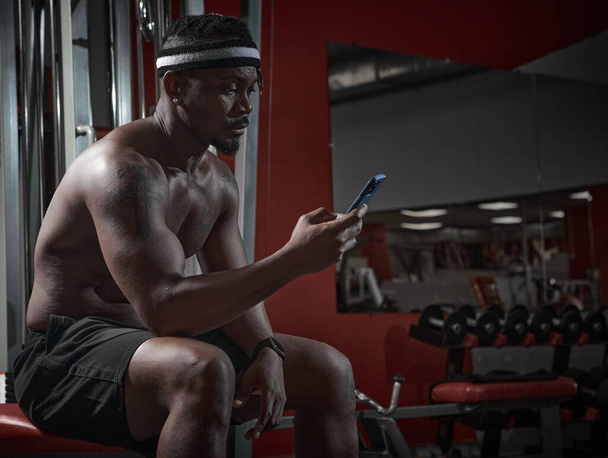 African american athletic man with phone using sports app for exercising and gym workout sitting on sport bench - Photo, Image
