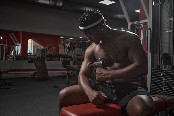 Athletic african american man massaging hand by handheld massage gun, post-workout recovery routines in gym - Foto, Imagem