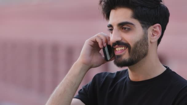Onnellinen hymyilevä parrakas intialainen, jolla on kännykkä korvassa puhumassa ystävän kanssa. Millennial mies opiskelija kommunikoi tyttöystävä. Mies keskustella liike, puhelut taksi tai tehdä tilauksen tavaroiden puhelimessa - Materiaali, video