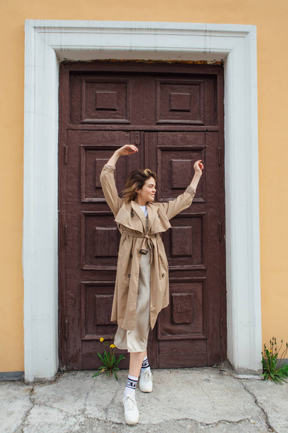 Young smiling millennial woman with wild hair dressed in an autumn coat posing near the door of an old building. - Фото, изображение