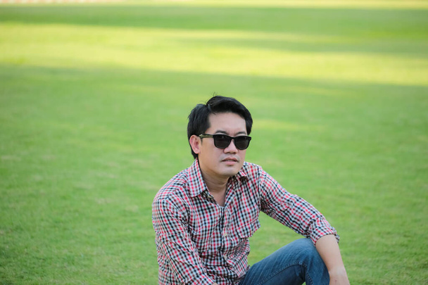 Homem vestindo camiseta Cheque, jeans azul sentado no gramado verde usando óculos escuros - Foto, Imagem