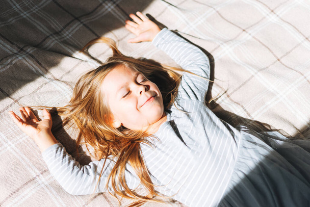Cuty funny little girl with long hair in grey home dress having fun lies in sun light on bed at home - Foto, afbeelding