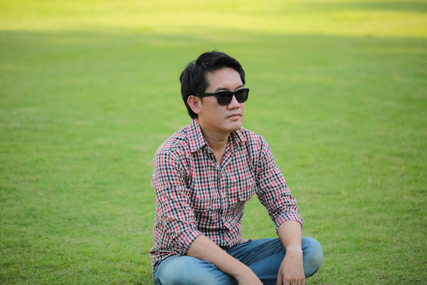 Чоловік у сорочці Check, сині джинси сидять на зеленому газоні в чорних сонцезахисних окулярах
 - Фото, зображення
