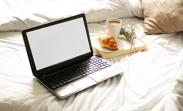 白いベッドの上にお茶が寝そべってるオープンノートパソコン。ベッドにはコーヒーとクロワッサンが入ったノートパソコンがあります. - 写真・画像