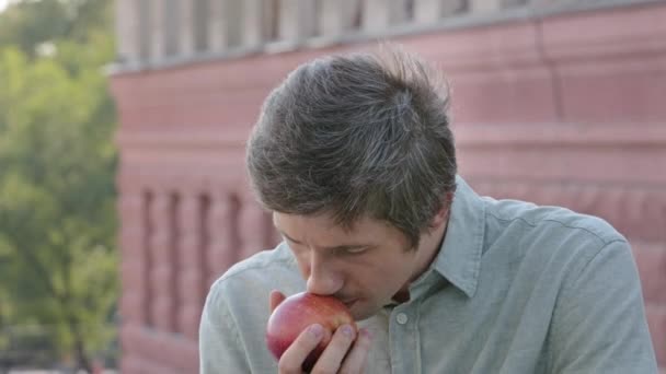 Young male vegetarian vegan holds beautiful ripe apple, inhales delicious aroma. Attractive dieting guy enjoying healthy vitamin breakfast or lunch, sniffing fragrant fruit, closing eyes admiringly - Footage, Video