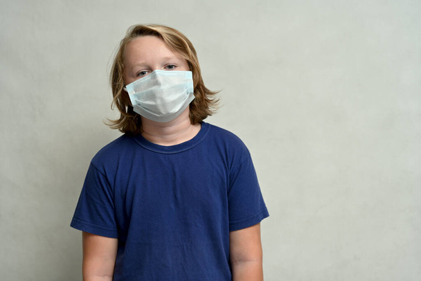 Young boy teenager in a medical mask with long hair in a blue t-shirt posing on a white background - Foto, Imagem
