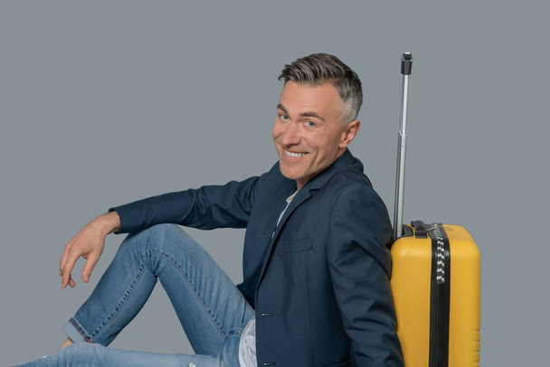 Joyful man sitting on floor near suitcase - Фото, изображение