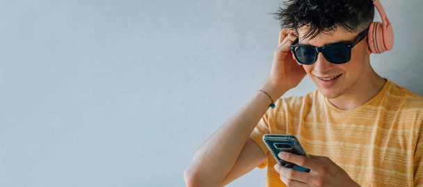 adolescente con gafas de sol, teléfono móvil y auriculares en fondo de pared gris - Foto, Imagen