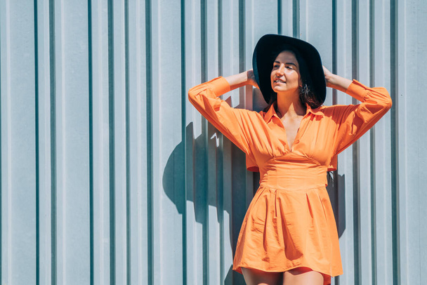 chica joven de moda en vestido naranja y sombrero en fondo de pared gris al aire libre - Foto, Imagen