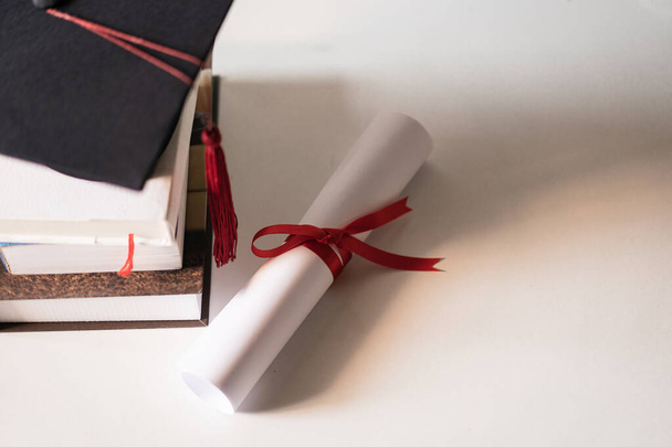 Maturitní klobouk nebo minometná deska a certifikát diplom papír svázaný červenou stuhou na hromadě knih s prázdným prostorem mírně nenasycený s dálniční známkou pro vintage efekt - Fotografie, Obrázek