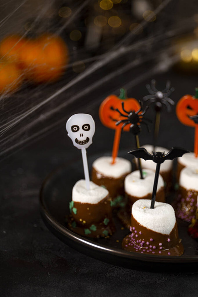  Sweet Halloween treat, chocolate covered marshmallows with sugar and Halloween decorations on a black plate, vertical photo with soft focus - Foto, imagen