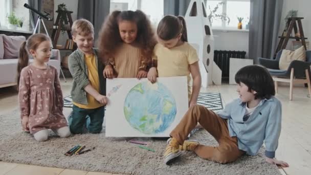 Portret grupy dzieciaków trzymających rysunek planety Ziemia i rakiety oraz pozujących do kamery w salonie - Materiał filmowy, wideo