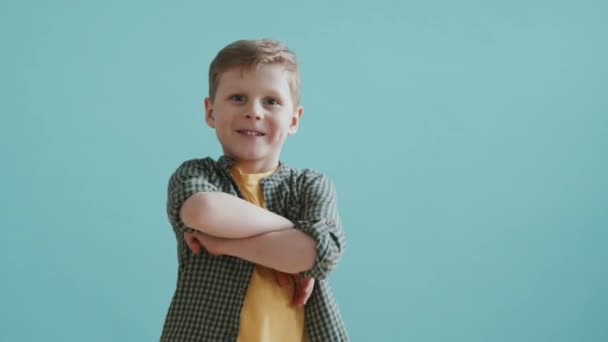 Portraitaufnahme eines süßen 6-jährigen Jungen, der seine Arme verschränkt und vor blauem Hintergrund für die Kamera lächelt - Filmmaterial, Video