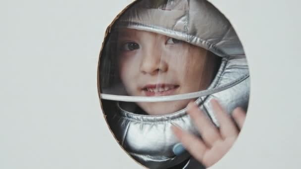 Kézzel tartott közeli felvétel aranyos kislány űrsisakban néz ki karton rakéta hajó ablak és integetett a kamerába - Felvétel, videó