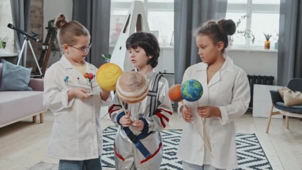 Handheld portret met slowmo van 8-jarige meisjes in labjassen en 6-jarige jongen in ruimtepak met zonnestelsel model en speelgoed planeten en chatten, dan poseren voor camera in de woonkamer - Video