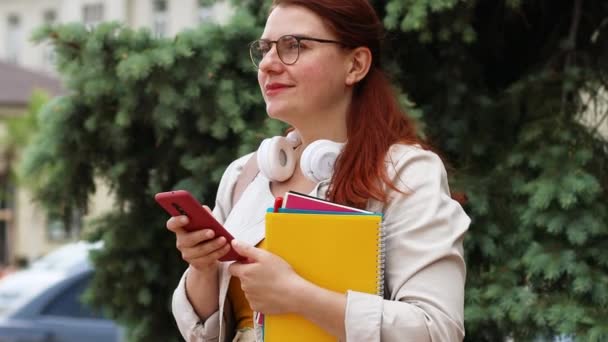 Osoba používá smartphone. Moderní technologie. Mladý dospívající dívka student v brýlích pro vidění a bezdrátové sluchátka s knihami a notebooky v městském parku. - Záběry, video
