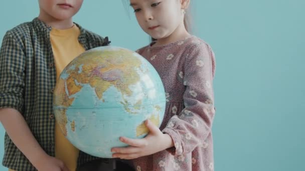 Incline-se retrato tiro de menino bonito 6-year-old e 7-year-old menina segurando globo mundial e sorrindo para a câmera enquanto posando contra fundo azul - Filmagem, Vídeo