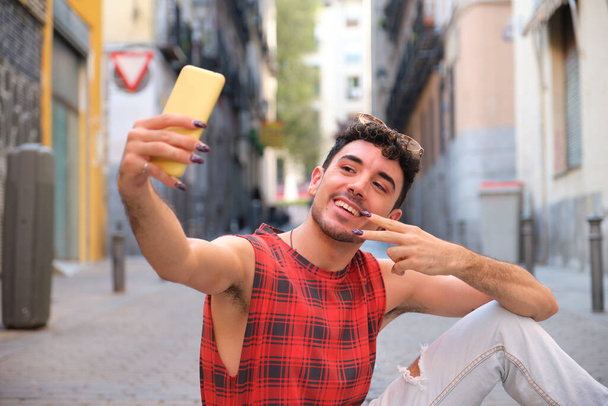 Joven hombre caucásico con uñas largas y falsas tomando una selfie con el teléfono inteligente sentado en el suelo. - Foto, Imagen