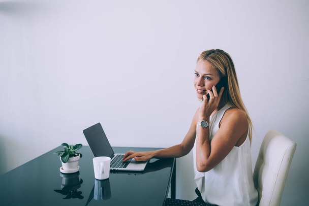 Side view of charming well dressed executive woman looking away and smiling while sitting at black glossy table with computer and mug of hot beverage and talking on phone against white wall in modern office - Foto, imagen