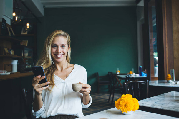 Cheerful relaxed woman with cup of fresh beverage looking at camera with smile while sitting at table with vivid yellow flowers and surfing smartphone in modern cafe - Φωτογραφία, εικόνα