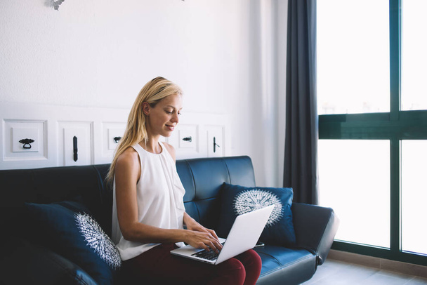 Side view of pleased blond haired woman in casual wear smiling while typing on keyboard of laptop in modern living room in Barcelona  - Photo, image