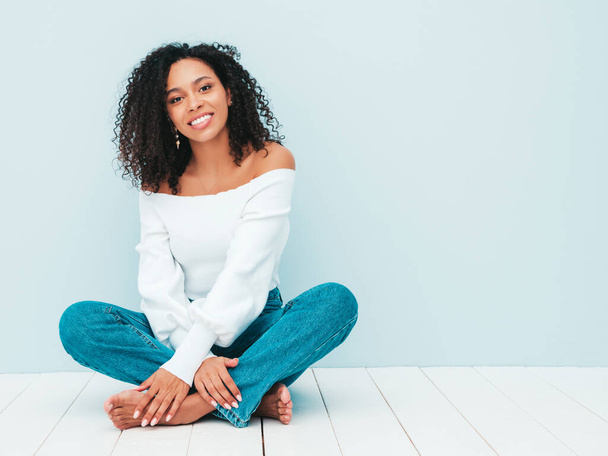 Bella donna nera con riccioli afro acconciatura.Modello sorridente in maglione e jeans alla moda. Sexy donna spensierata seduta vicino alla parete azzurra in studio. Abbronzato e allegro - Foto, immagini