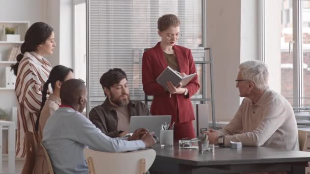 赤の正式なスーツを着た白人女性会計士の中長期,ビジネスミーティングにメモを取ります,会議テーブルのそばに立って,多様な同僚が座って - 映像、動画