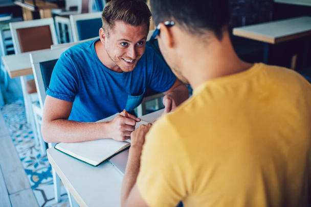 Cheerful caucasian male journalist writing notes during conversation interview with man in coffee shop satisfied with job and friendly talk, positive hipster guys cooperating making research togethe - Foto, Imagem