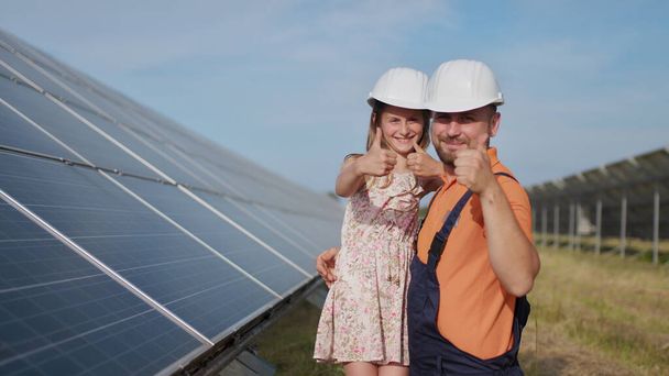 Батько, інженер-сонячна енергетика, і його дочка стоять біля сонячних панелей. Батько пояснює дитині принцип сонячної електрики і кладе захисний шолом на голову дівчат
. - Фото, зображення