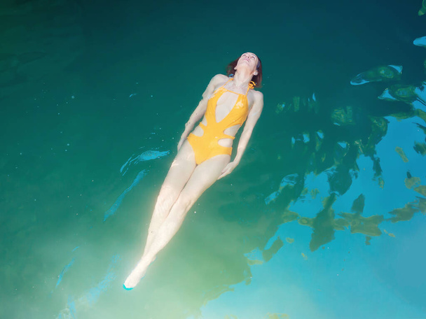 Een meisje in een geel zwempak strekte zich uit over haar rug in een schone turquoise vijver. - Foto, afbeelding