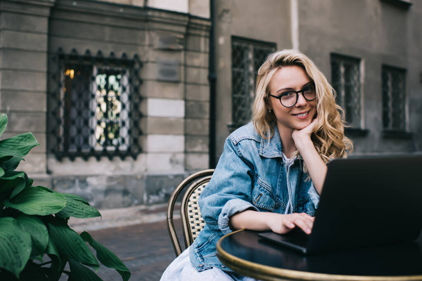 Beautiful young female in denim jacket and glasses smiling and looking at camera while using laptop and sitting at cafe table on city street - Фото, изображение
