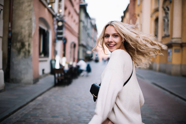 Back view of excited romantic young blonde in casual outfit and with belt bag spinning around on street and looking over shoulder at camera  - Valokuva, kuva