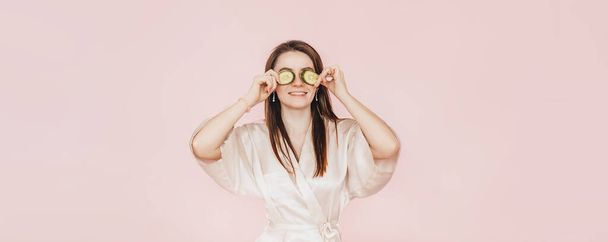 Girl make homemade face beauty masks. Cucumbers for the freshness of the skin around the eyes. Woman take care of youthful skin. Model laughing and having fun in spa on pink background - Valokuva, kuva