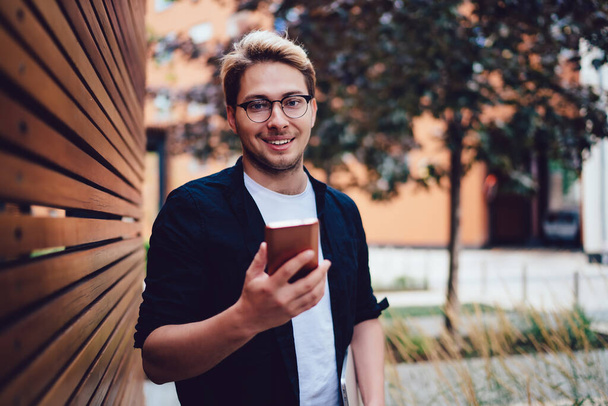 Joyful young male in casual wear and glasses using cellphone messaging with colleague spending time in street looking at camera - Foto, Imagem