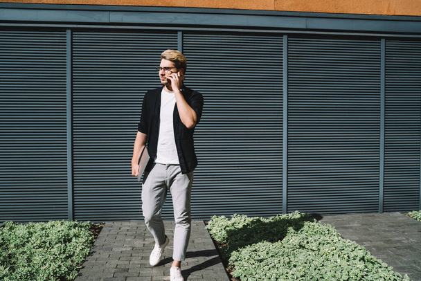 Young serious male in glasses dressed in grey pants black jacket and white t shirt having conversation on mobile phone while walking on street at daytime - Zdjęcie, obraz