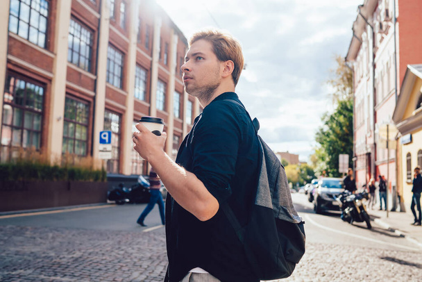 From below of careful young man in casual wear with backpack taking cup of coffee traveling in city looking away in back lit - Фото, зображення