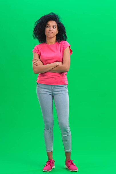 Portrait of young female African American thinking about something, upset that there is no idea. Black woman with curly hair in pink tshirt and jeans poses on green screen in the studio. Full length. - Foto, Imagem