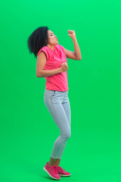 Portrait of young female African American cheerfully dancing the dance of victory, joy or good mood. Black woman with curly hair poses on green screen in studio. Full length. - Foto, Imagem