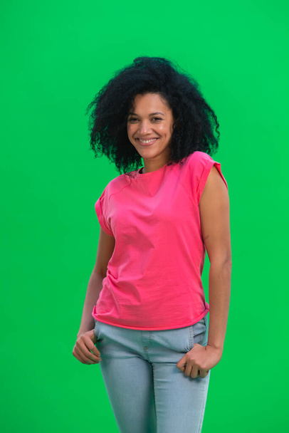 Portrait of young female African American smiling happily. Black woman with curly hair in pink tshirt poses on green screen in the studio. Close up. - Fotoğraf, Görsel