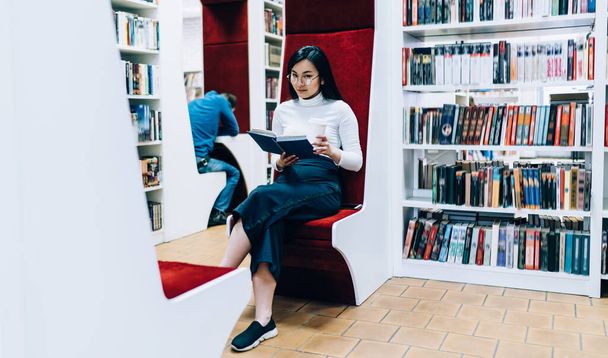 Young Asian woman drinking coffee and looking at camera while sitting on comfortable red chair and reading by faceless man in book shop - Foto, immagini