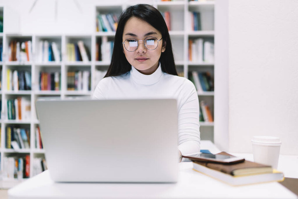 Mujer adulta asiática pensativa en gafas y ropa formal que se concentra en la pantalla e interactúa con el ordenador portátil mientras está sentado en el escritorio con el cuaderno de notas del teléfono y tomar una copa en la biblioteca en el fondo borroso de las estanterías - Foto, imagen
