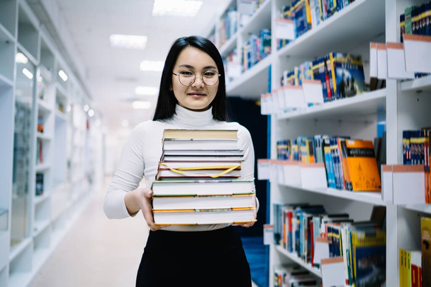 Dark haired female with glasses in white turtleneck and black trousers holding books in modern bright library while looking at camera - Foto, immagini
