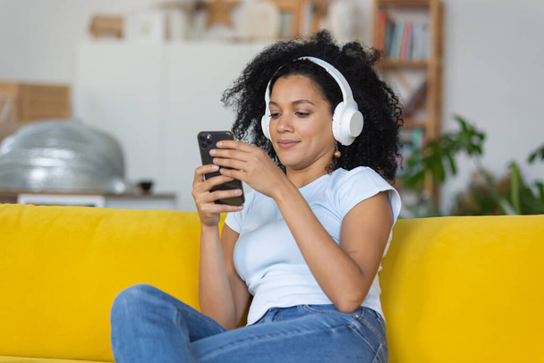 Portrait of a young African American woman enjoys music with big white headphones using smartphone. Brunette with curly hair sitting on yellow sofa in a bright home room. Close up. - Fotoğraf, Görsel