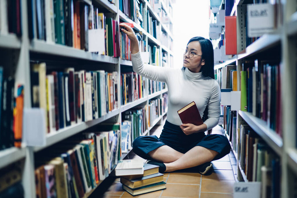 Modern Asian woman sitting between bookshelves in university library with stack of books on floor and taking books for preparing to exam - Φωτογραφία, εικόνα