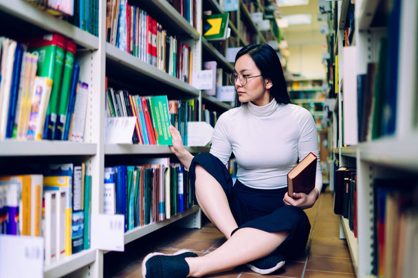 Serious young Asian female student sitting on floor next to bookshelf and looking through books while searching information for study - Foto, Bild
