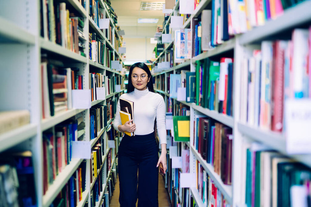 Dark haired Asian female with glasses in white turtleneck and blue trousers holding colorful books and walking between bookcases in library - Photo, image