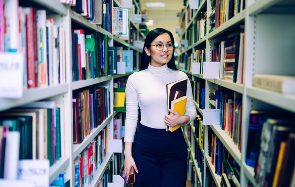 Dark haired young Asian woman in round glasses with colorful notebooks and smartphone smiling and walking between bookcases in library - Foto, Imagem